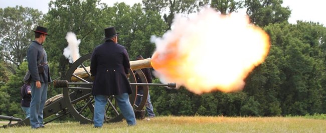 Three men in Union Civil War outfits firing a cannon in a field with a tree line behind them