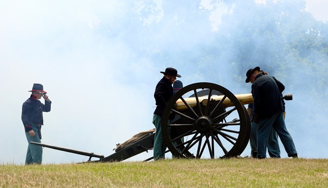 An artillery crew in blue uniform mans a bronze cannon while smoke fills the air