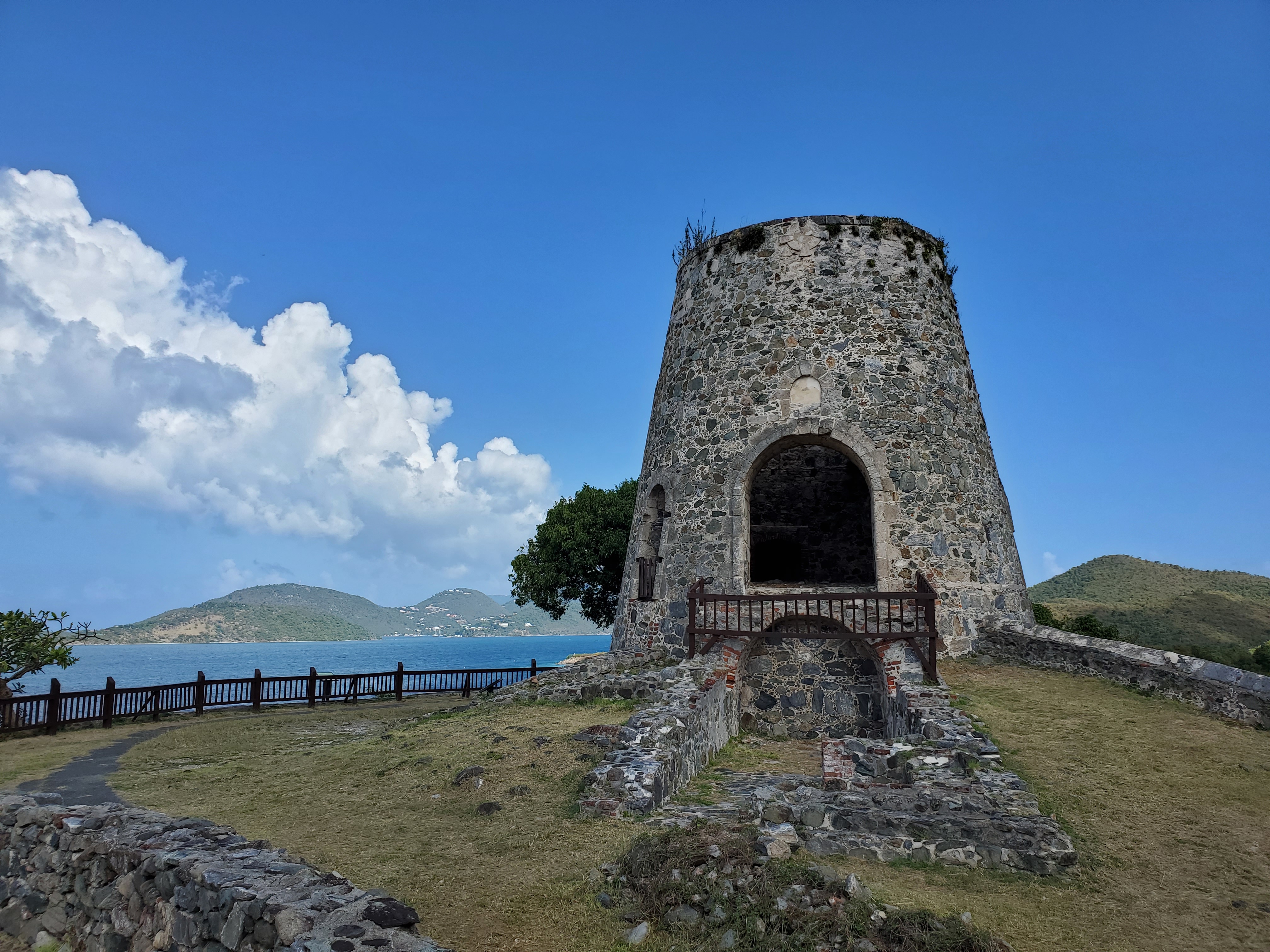 Photograph of Annaberg Windmill atop a grassy hill with ocean and blue sky in the background.