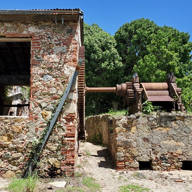 Large, rusted horizontal rollers connect through a rusted shaft to a large gear attached to a brick, stone, and coral masonry building