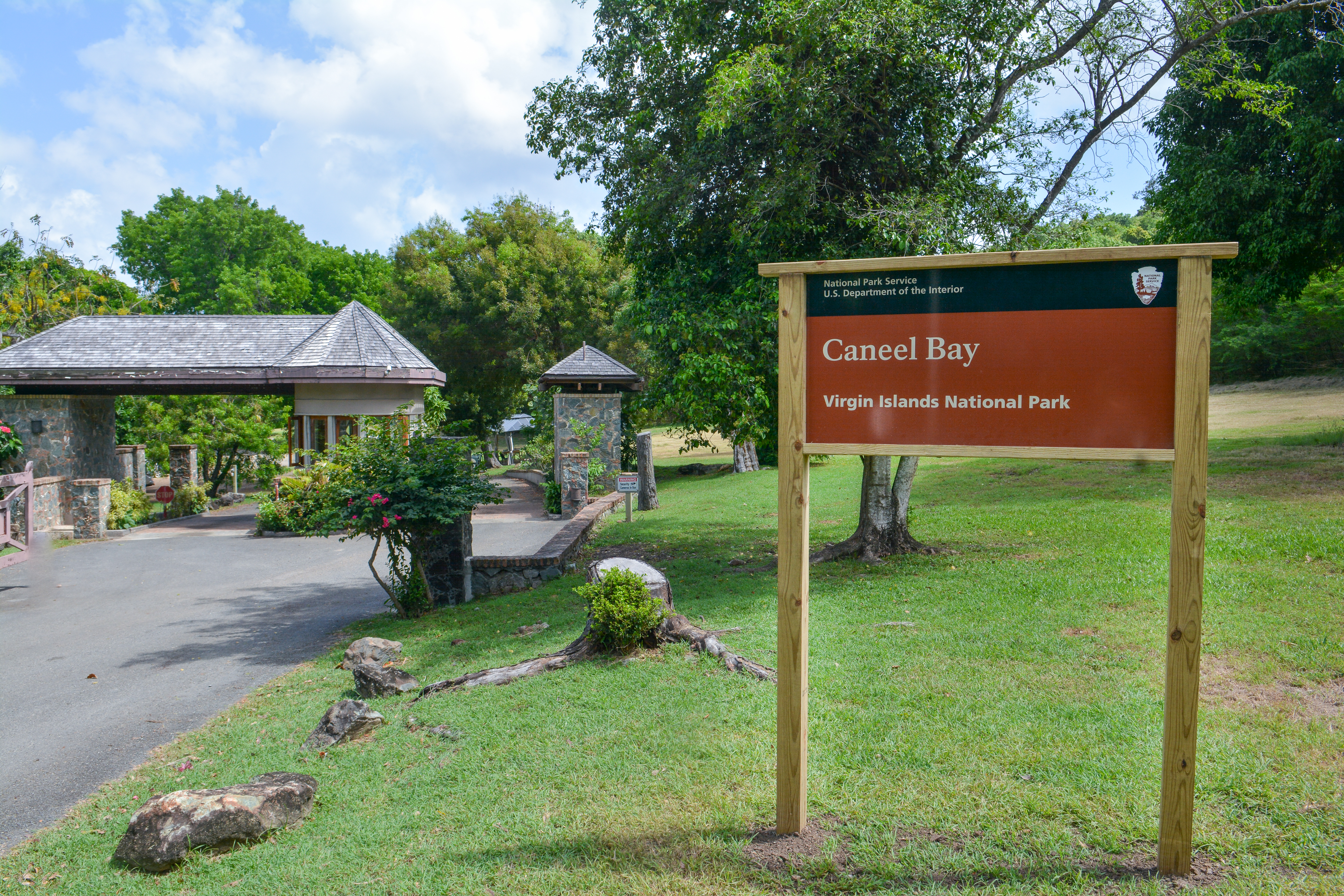 Image of a sign reading Caneel Bay in front of a driveway and small builing with green trees and blue sky.