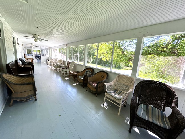 A long row of wooden and wicker chairs line the sides of a long screened-in porch on a bright summer day.