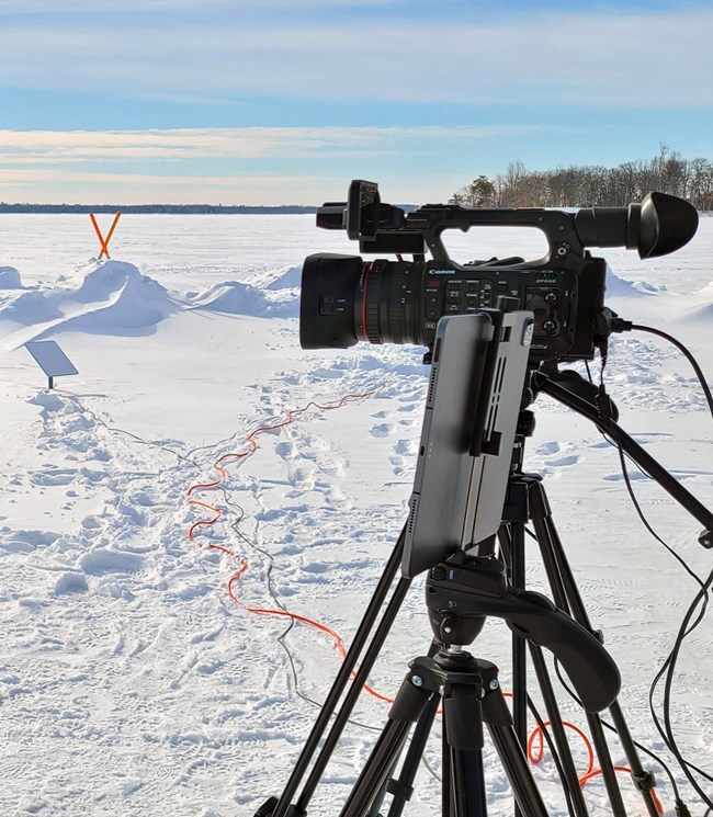 A large camcorder and tablet--both mounted to tripods--point towards a frozen scenic lake. Wires and extension cords run past the cameras into the snow.