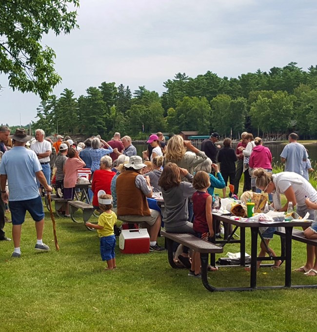 A large group mingles and dines at a grassy picnic area on the shores of Lake Kabetogama