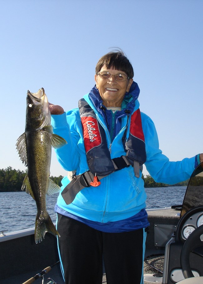 A woman stands in a boat and holds a recently-caught walleye up for the camera.