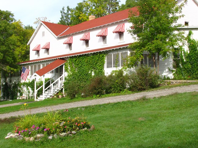 A large, two-story building with a red roof and red awnings sits at the top of a green hill, with a small flower garden in the front.