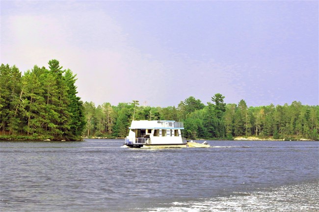 A white and blue houseboat tows a metal fishing boat behind it along the surface of a tree-lined scenic lake.