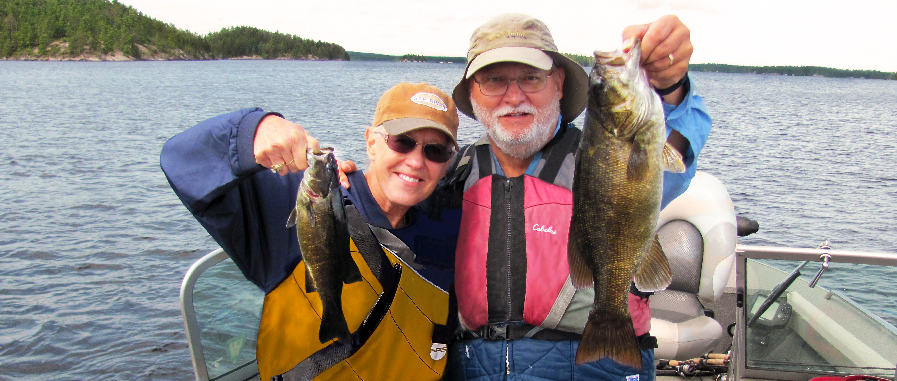 A husband and wife stand in a boat, each holding up a smallmouth bass.