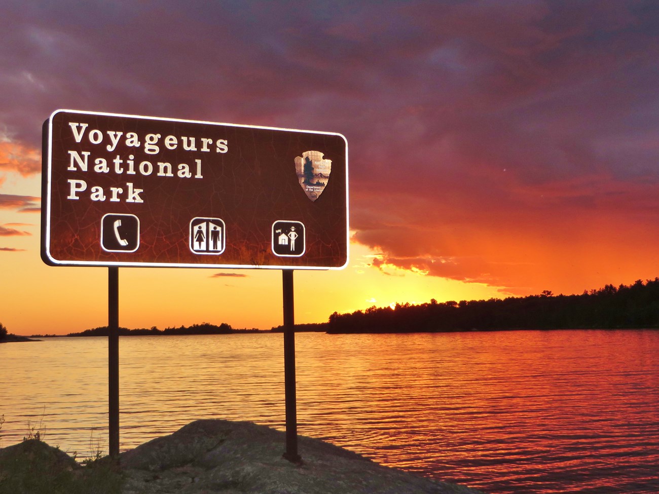 A scenic, tree-lined lake reflects a vivid orange, red, and yellow skyline at dusk, with a large Voyageurs National Park sign in the foreground.