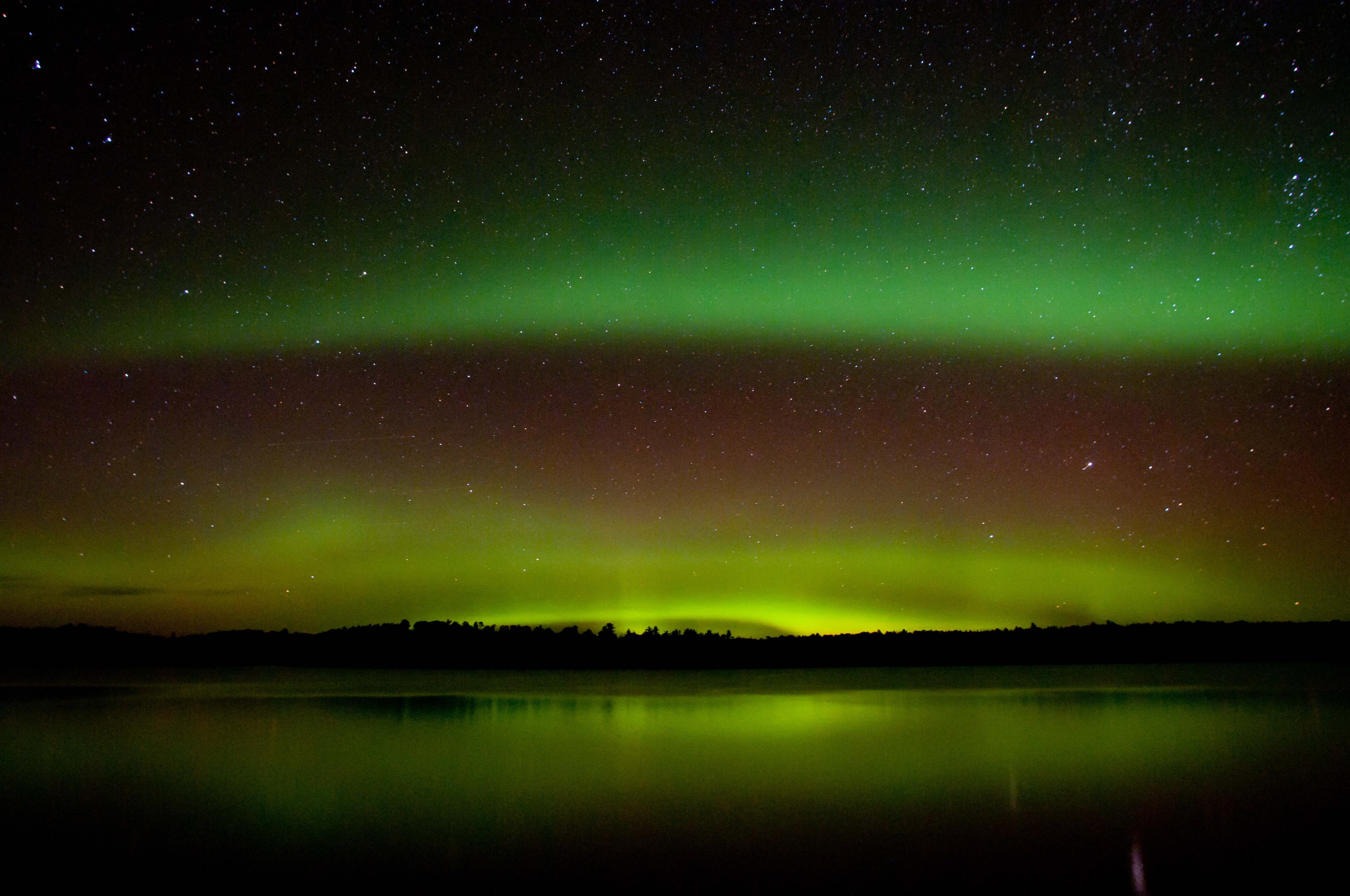 Vivid, wavy bands of green and red light shine against a dark sky over a scenic lake