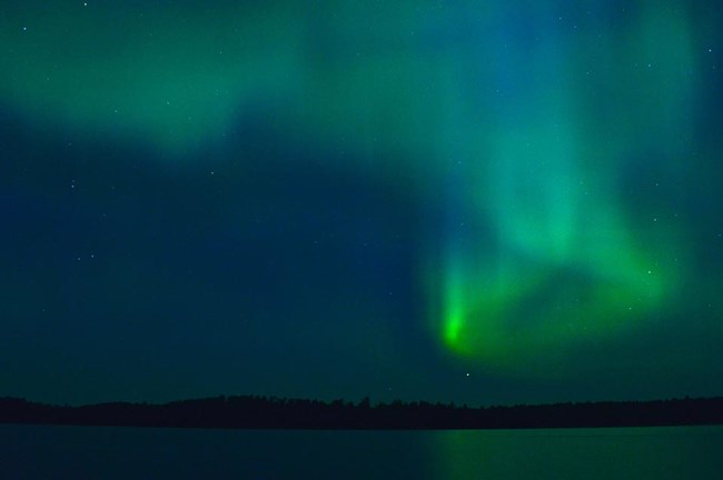 Bands of green and blue light appear to dance and swirl in the dark night sky above a scenic lake.