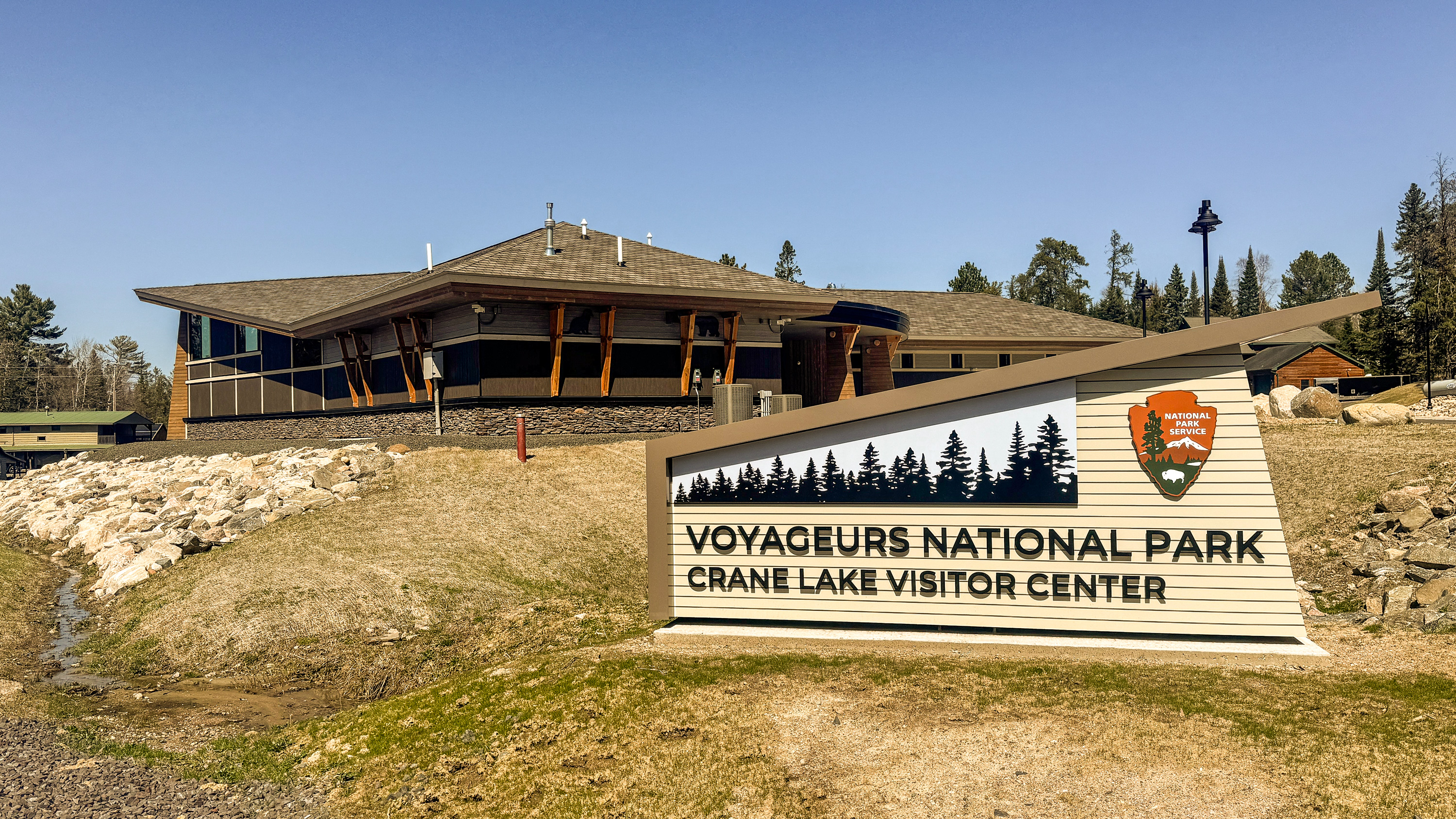 New Crane Lake Visitor Center sign with building in the background on a sunny spring day with the ground still brown and no grass yet.