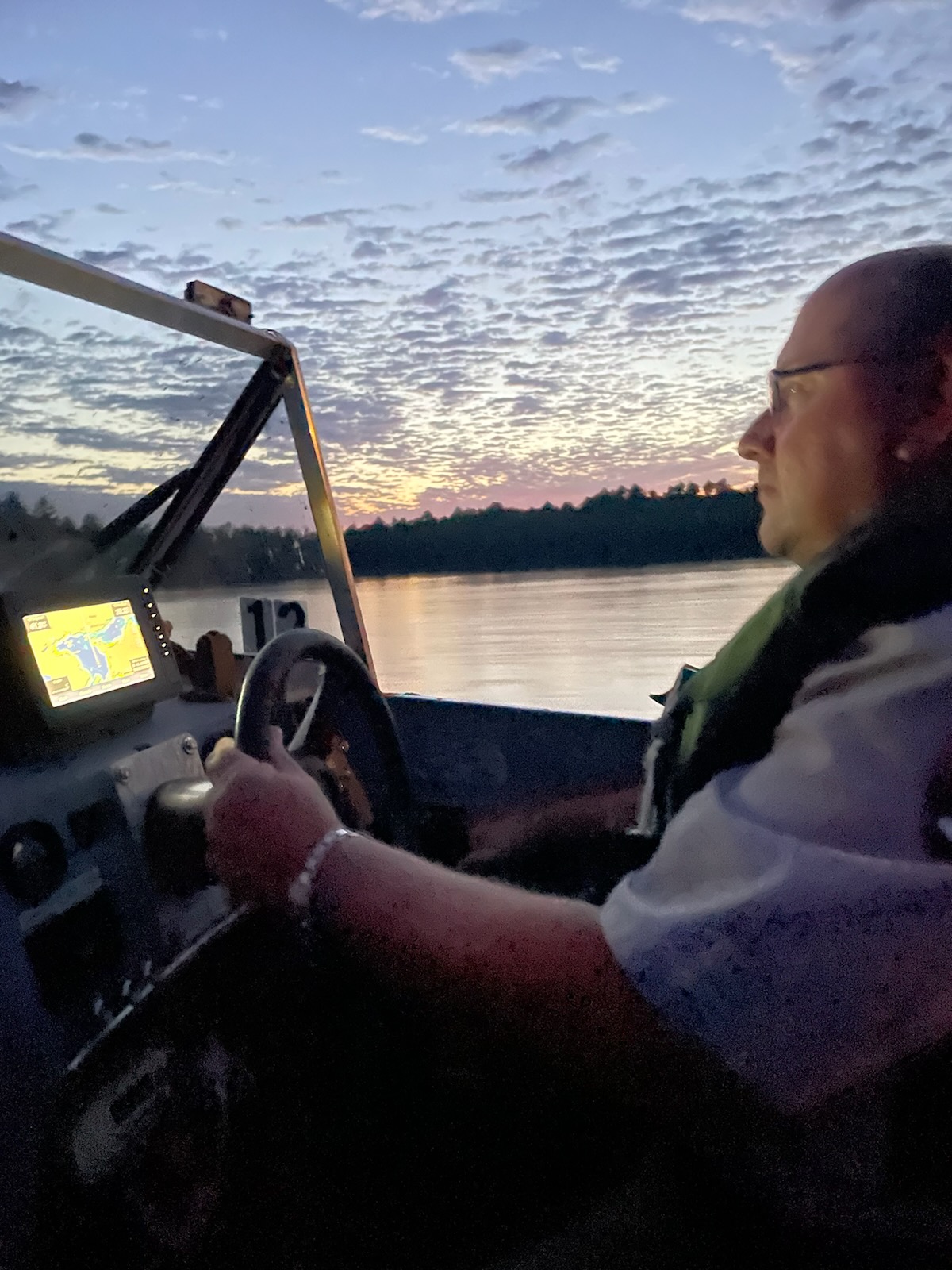 Ranger wearing a personal flotation device behind the wheel of a motor boat. The sun sets in the background against scattered clouds.