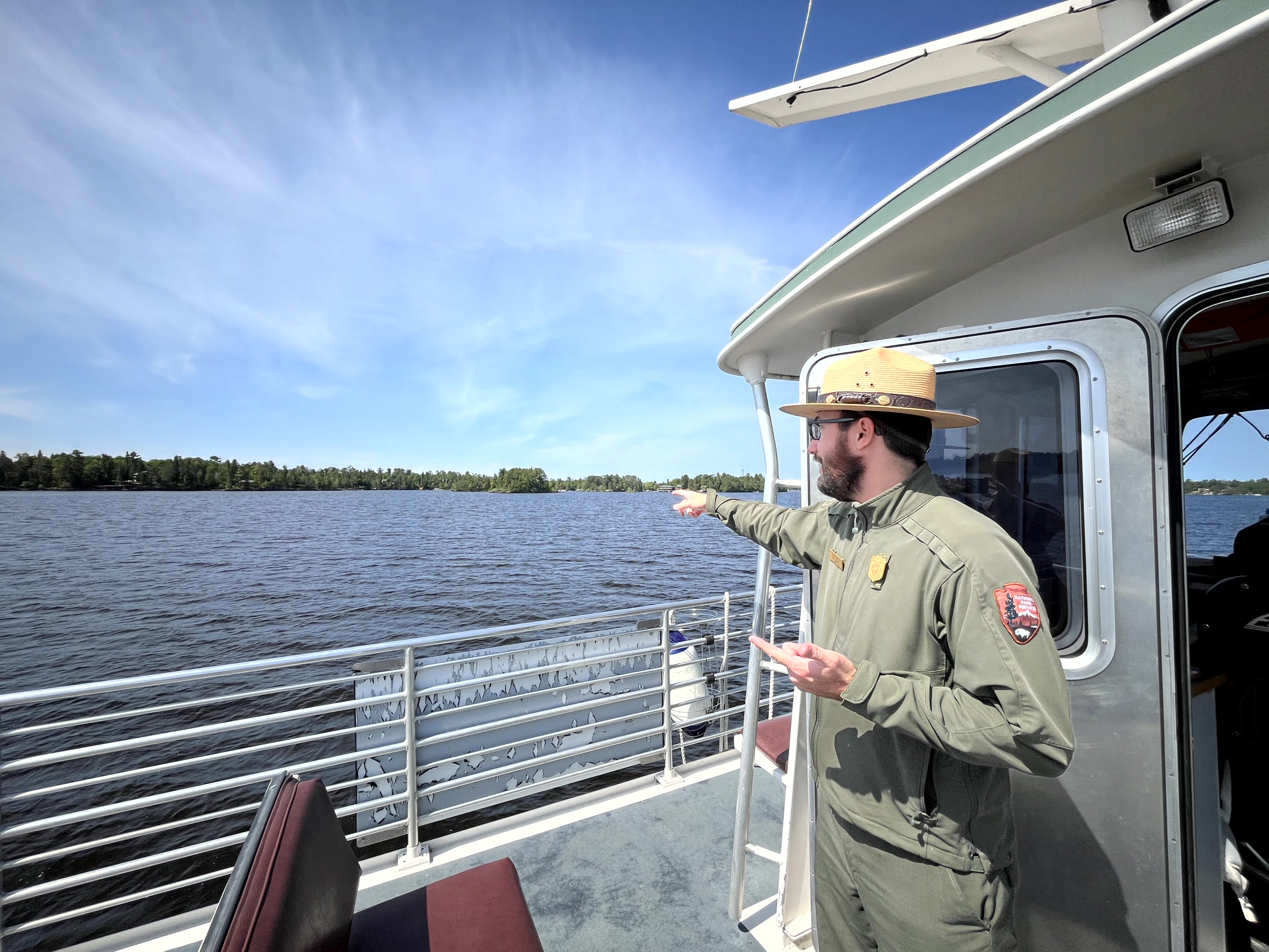 A Park Ranger in NPS uniform is standing on a boat and pointing across the waters of the lake at the forested shoreline. The sky is blue and partially cloudy.