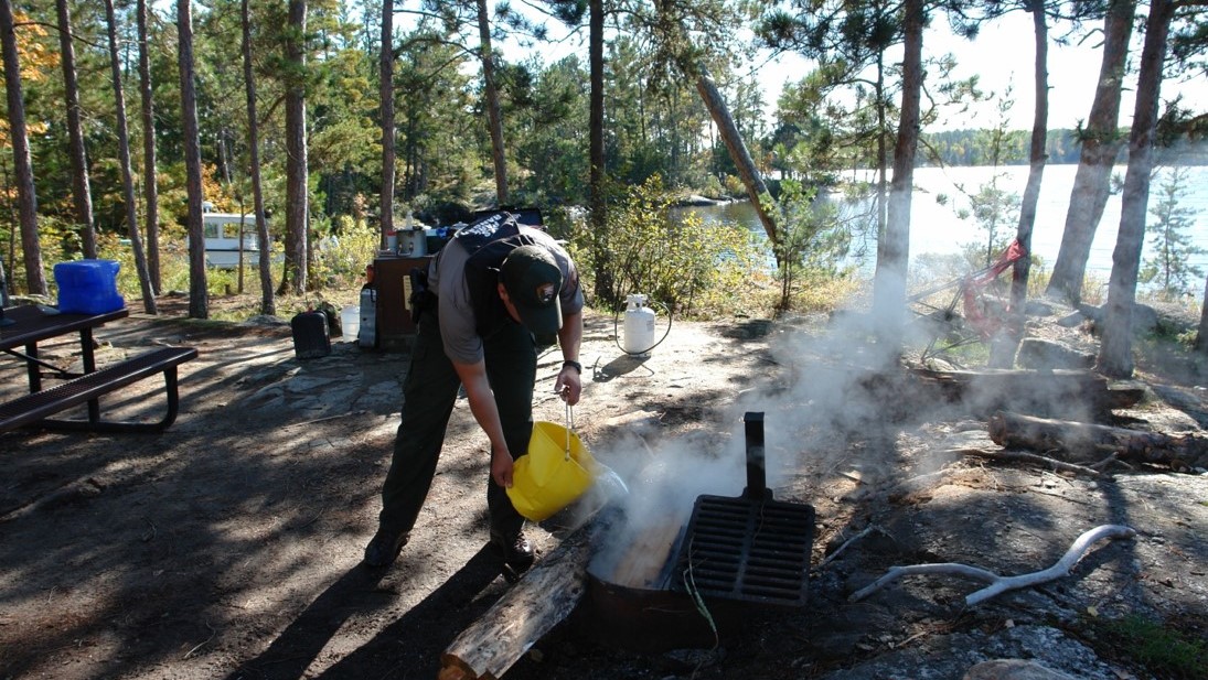 A Park Ranger uses a bucket of water to put out a campfire.