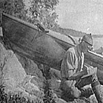 Historic photo, man sitting on rocky shoreline writing in a journal with boat pulled up next to him.
