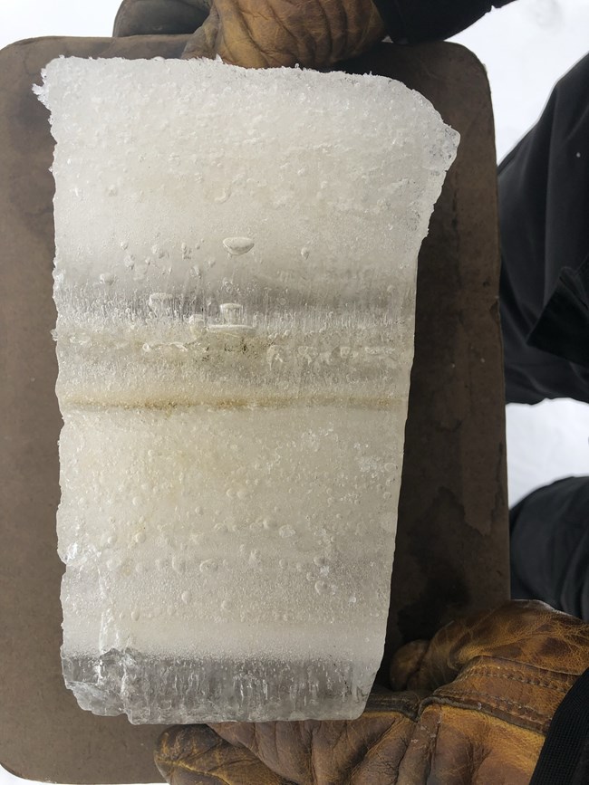 Ice core sample held by two gloved hands.  Alternating layers of white ice and clear ice thoughout 8 inches of total ice.