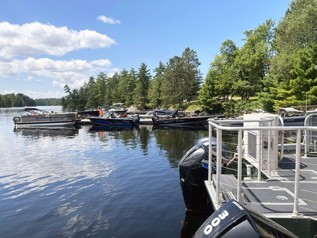 Groups of motorboats cluster along a series of docks on a scenic shoreline.