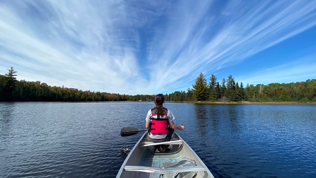 A young woman wearing a red life jacket paddles a metal canoe across the waters of a scenic lake.