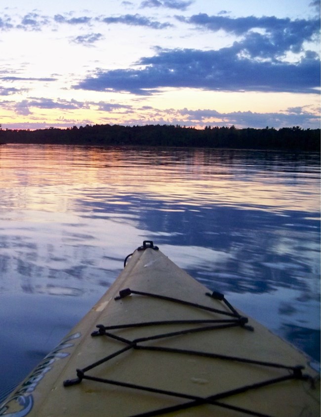 The nose of a yellow kayak points across the still waters of a scenic lake, reflecting a purple and gold sunset.