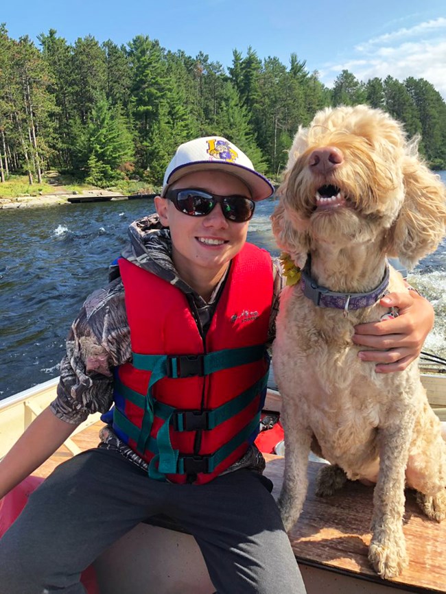 A boy in sunglasses sits smiling in the back of a motorboat with his arm around a large, shaggy, tan dog.