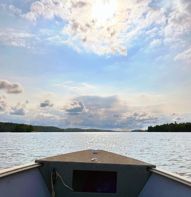 The nose of a boat points towards the bright, sunlit horizon of a scenic lake.