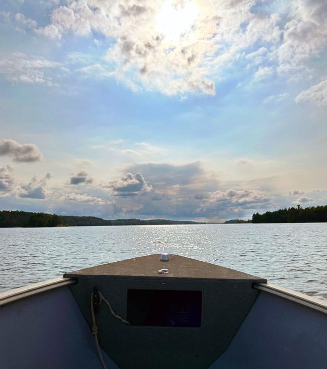 The open bow of a metal boat faces towards a scenic lakeshore surrounded by green trees.