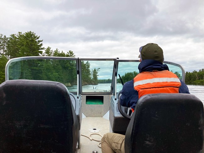 A woman drives a motorboat through a narrow area rimmed with evergreen trees on a scenic lake