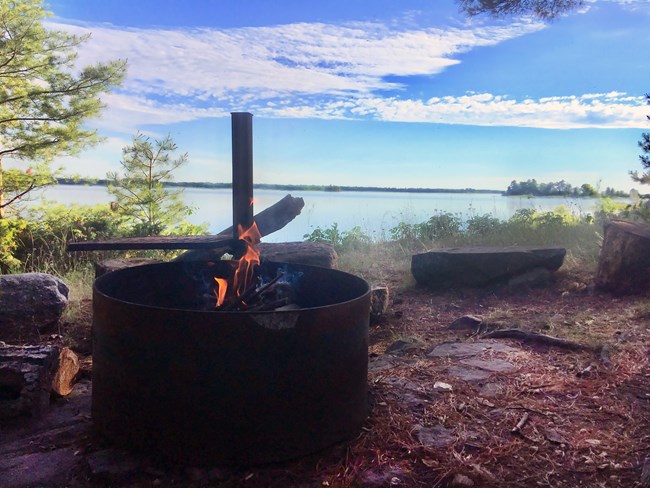 A campfire burns in a grate overlooking a campsite on a scenic lake.