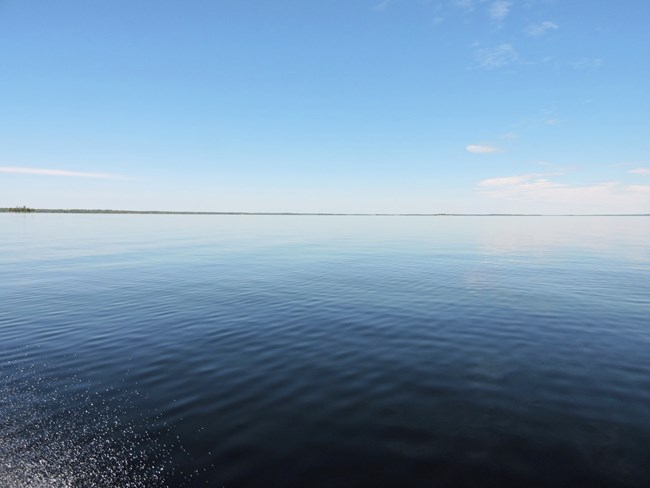 A spray of boat wake on a vast, blue lake with a band of small trees on the far horizon.