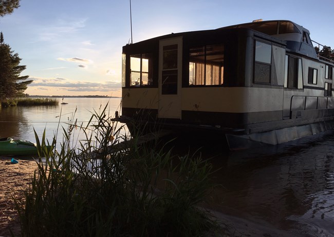 The rear of a large houseboat connects to a warm, light-filled beach by way of a ramp.