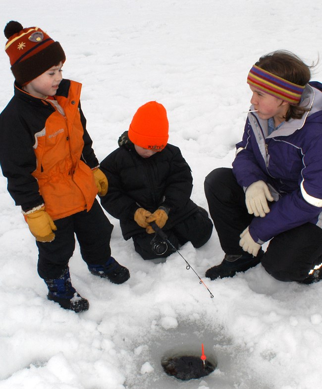 Three children in colorful winter clothing huddle around a round hole in the ice on a frozen lake. One child holds a small fishing pole with a bobber attached.