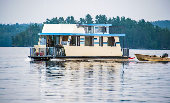 A large, boxy yellow-white houseboat glides across a scenic lake with pine trees on the shoreline.