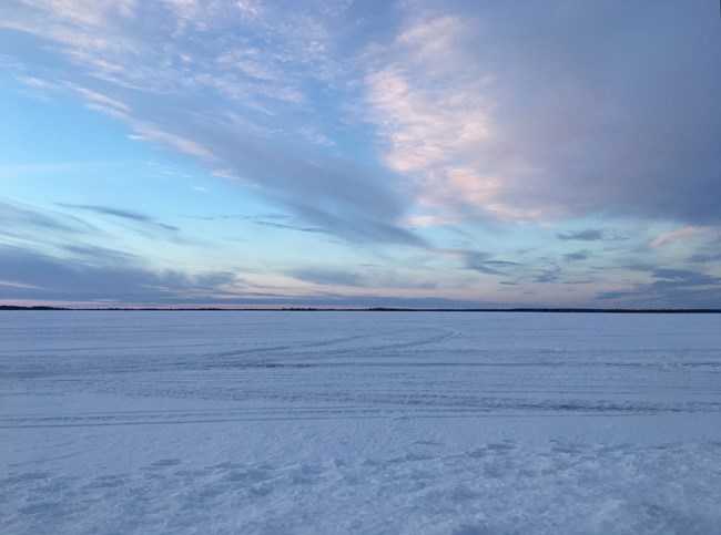 The frozen surface of a scenic lake is tinted to pastel blue and pink hues as the sun sets.