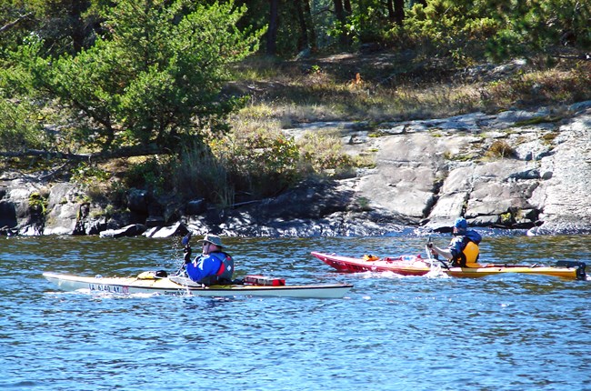 Two adults in life jackets paddle brightly-colored kayaks along a scenic rocky shoreline dotted with shrubs and trees.