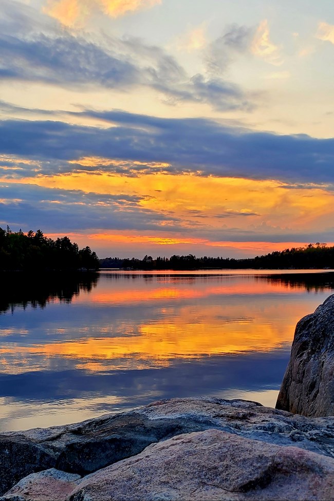 A colorful red, blue, gold, and purple sunset reflects on the calm waters of a large, scenic lake.