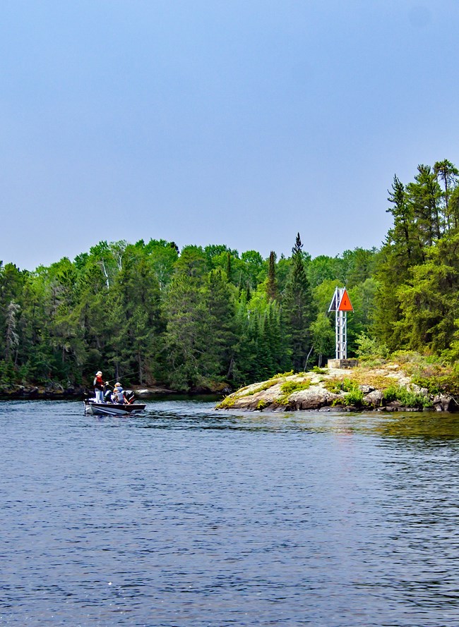 Three people sit in a black and white boat and fish near the rocky shoreline of a scenic lake.