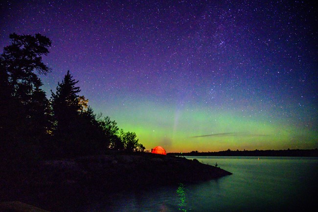 A single red tent sits on a tree-lined lakeshore, illuminated from behind by a band of green and yellow northern lights.