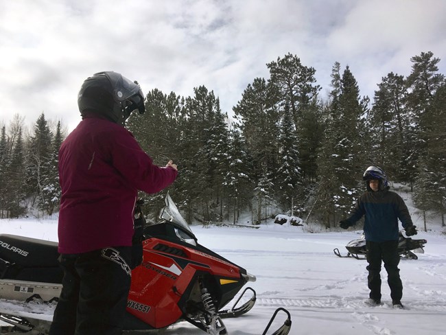 Two snowmobilers dressed in heavy winter gear give each other the "thumbs up" signal at the edge of a scenic winter trail through the woods.