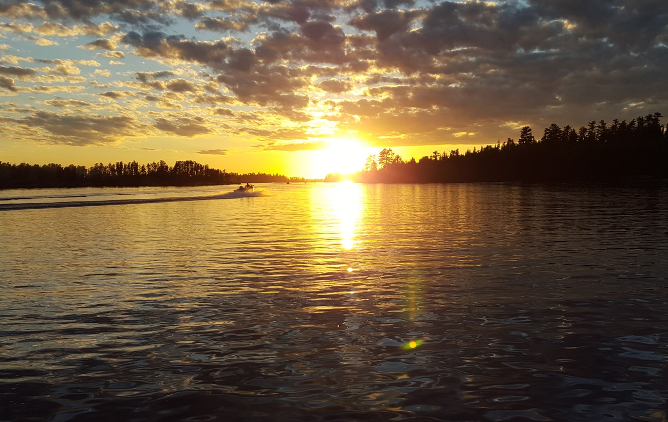 Several silhouetted motorboats drive towards the horizon of a scenic lake as the sun sets.