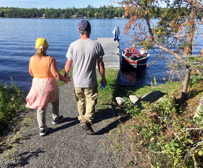 A man and woman walk hand in hand towards a boat docked on the shore of a scenic lake