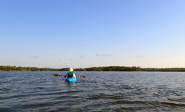 A kayaker in a blue craft paddles on a large, scenic lake with trees on the distant horizon