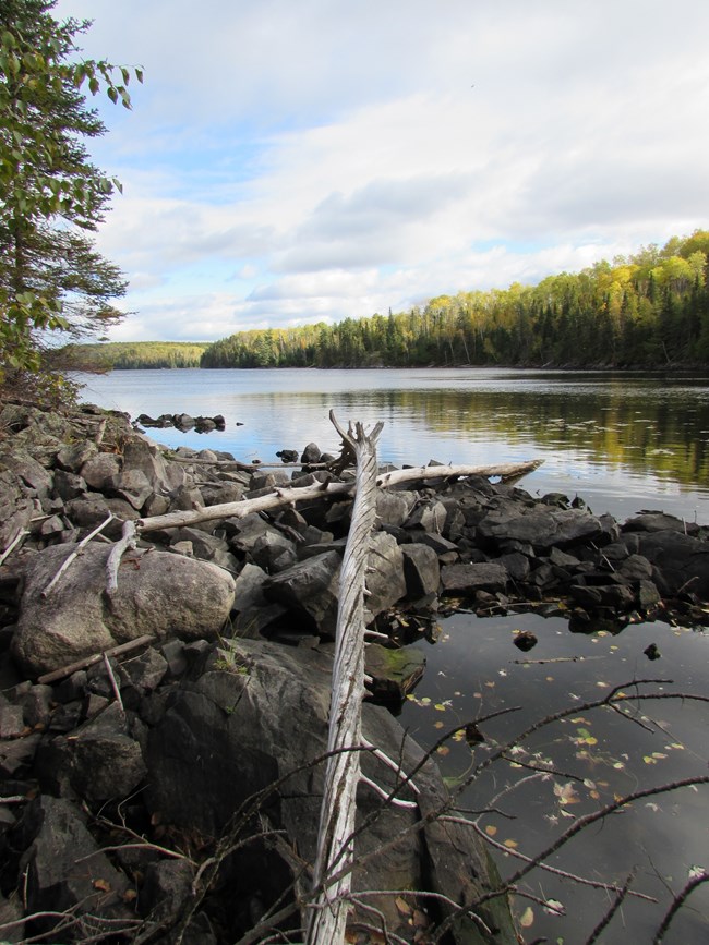 A calm lakeshore reflects surrounding rock ledges and trees.