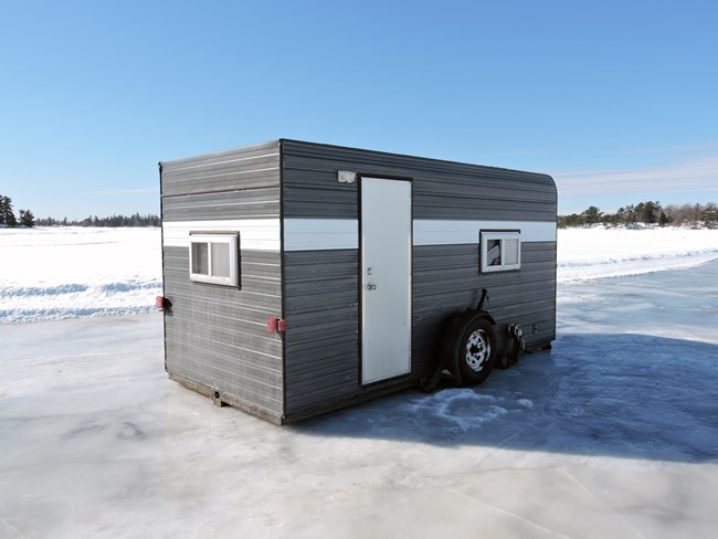 A large, boxy, metal ice fishing shelter sits on the surface of a frozen lake.