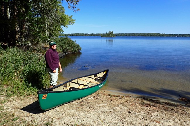 A woman stands on a scenic lakeshore next to a green canoe.