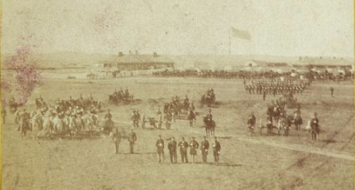 Fort Harker, Kansas. From 1860's photograph, shows soldiers posing in the foreground. In background is a barracks with the American flag overhead. Setting is on the Great Plains.