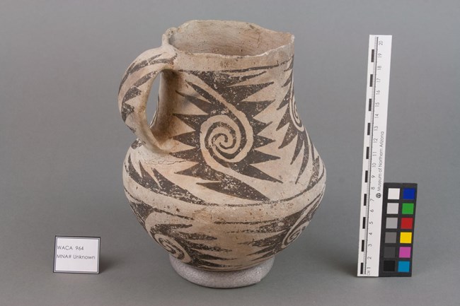 An ancient ceramic pitcher made of white clay with angular, black designs painted onto it. The pitcher is in a sterile setting with a label identifying it as "WACA 964, MNA # Unknown." Beside it are a series of color swatches and a ruler.