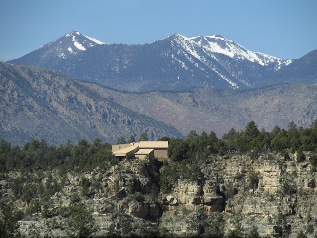 A visitor center sits across the canyon at cliffs edge surrounded by pine trees. The San Francisco peaks mountain range rises out of the horizon, dwarfing the canyon in comparison.