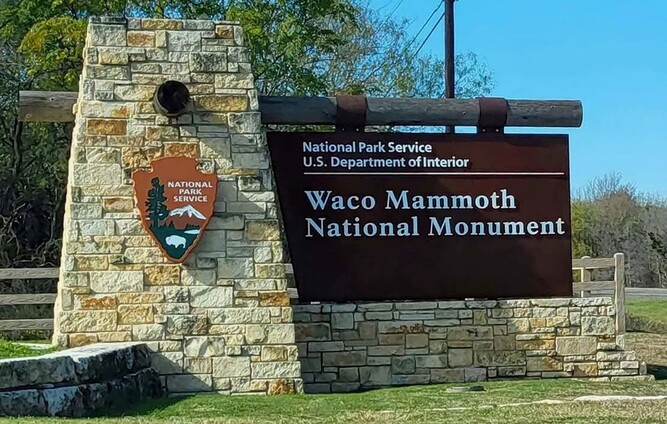 Brown Waco Mammoth National Monument sign with green grass, trees, and blue sky.