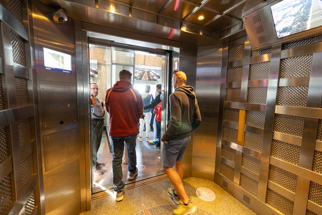 Elevator riders exit the elevator to land on the observation level while a ranger directs them out holding the door open. The elevator has a metallic interior and TV screen atop the wall.